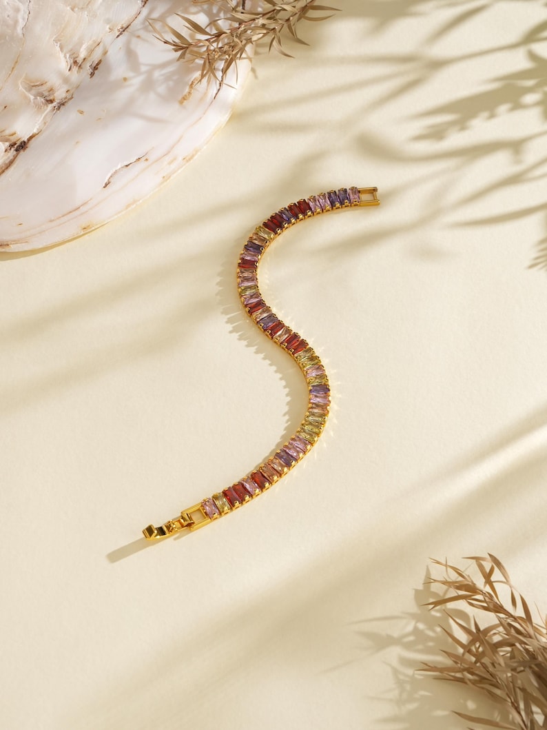 May include: A gold bracelet featuring a line of multi-coloured rectangular gemstones. The gemstones are in shades of red, purple, yellow, and green. The bracelet is on a cream surface with shadows and a seashell.