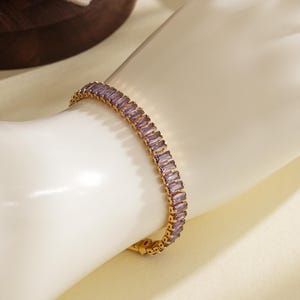 May include: Gold-toned bracelet featuring rectangular, light purple gemstones. The bracelet is set in a gold metal band and is displayed on a light-colored surface. The jewelry piece is a classic and elegant accessory.
