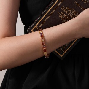 May include: A gold bracelet featuring rectangular, multi-coloured gemstones. The bracelet is worn on a wrist, and the person is holding a dark brown book with gold trim and the text "YOUR TEXT SAMPLE".