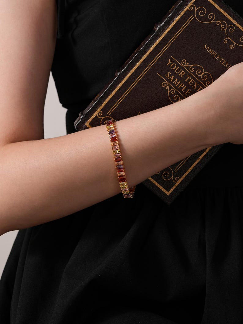 May include: A gold bracelet featuring rectangular, multi-coloured gemstones. The bracelet is worn on a wrist, and the person is holding a dark brown book with gold trim and the text "YOUR TEXT SAMPLE".