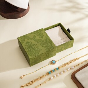 May include: An open green jewellery box with a patterned design, next to three gold bracelets. One bracelet has a turquoise stone, another has turquoise beads, and the third has pearls. A white jewellery display is in the background.