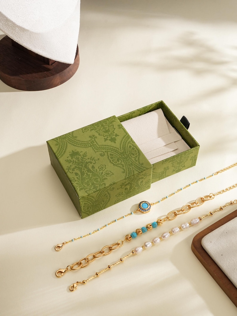 May include: An open green jewellery box with a patterned design, next to three gold bracelets. One bracelet has a turquoise stone, another has turquoise beads, and the third has pearls. A white jewellery display is in the background.