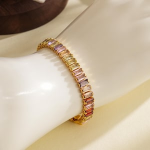 May include: A gold bracelet featuring rectangular, multi-coloured gemstones. The gemstones are arranged in a row, with colours ranging from purple to red. The bracelet is on a white surface.