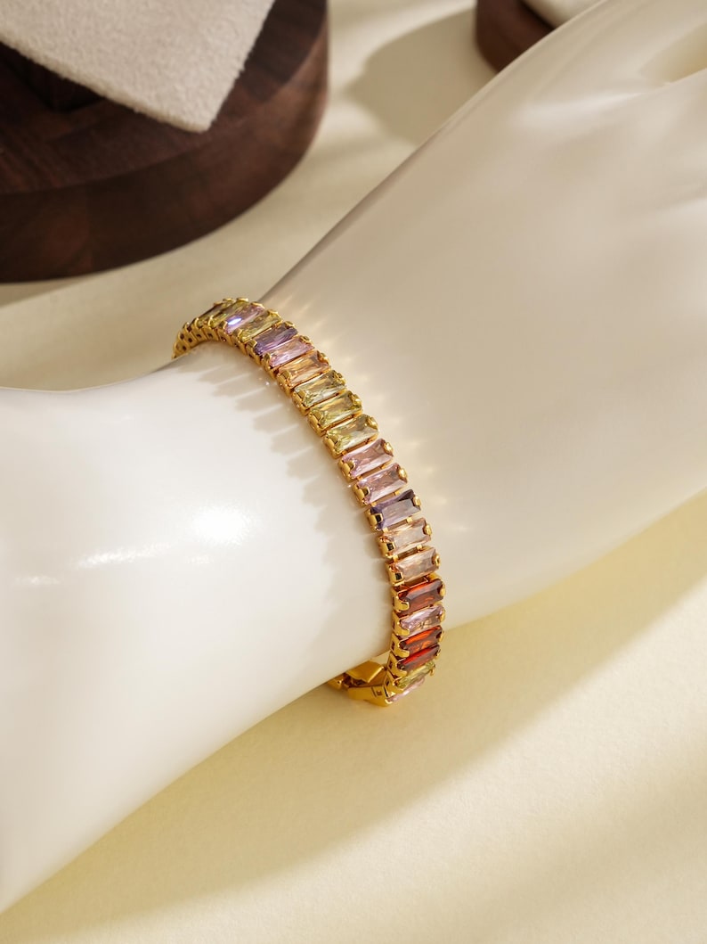 May include: A gold bracelet featuring rectangular, multi-coloured gemstones. The gemstones are arranged in a row, with colours ranging from purple to red. The bracelet is on a white surface.
