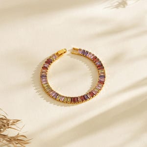 May include: Gold-plated bracelet with a series of multi-coloured rectangular gemstones. The bracelet is open, revealing the clasp and the array of purple, red, yellow, and green stones. The jewellery is set against a neutral background.