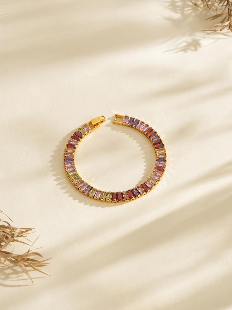 May include: Gold-plated bracelet with a series of multi-coloured rectangular gemstones. The bracelet is open, revealing the clasp and the array of purple, red, yellow, and green stones. The jewellery is set against a neutral background.