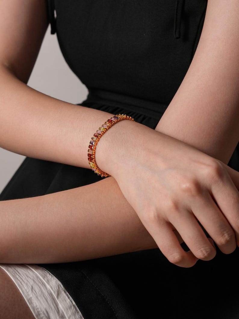 May include: A gold bracelet set with a row of colourful gemstones in shades of red, orange, yellow, and purple. The bracelet is worn on a person's wrist, with the arm and part of the black dress visible in the image.