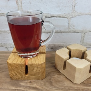 Könnte beinhalten: Ein Holz-Teebecherwärmer mit einem Glas-Teebecher gefüllt mit rotem Tee. Der Wärmer hat einen quadratischen Sockel und vier Schlitze, in denen der Becher ruhen kann. Es gibt einen separaten Holz-Kerzenhalter mit einer runden Öffnung für eine Kerze.