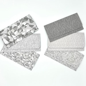May include: Six different patterned envelopes in shades of gray. The envelopes are arranged in two stacks of three. The patterns include geometric shapes, leaves, and dots.