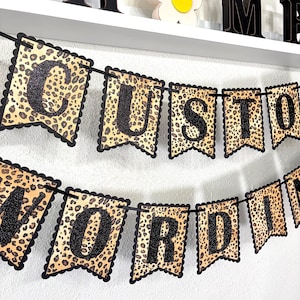May include: A decorative banner featuring scalloped-edge pennants with a tan and brown leopard print pattern. Each pennant displays a large, uppercase black glitter letter, spelling out the words CUSTOM WORDING strung on a black cord.