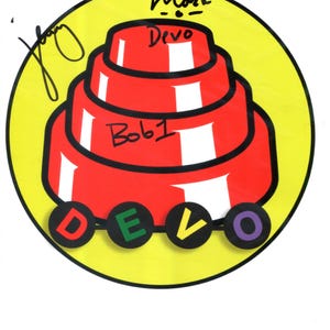 以下が含まれることがあります： 赤色の多層構造、単語「Devo」、および「Mark」と「Bob 1」の名前が特徴の円形グラフィック。文字D、E、V、Oは個別の円で表示されます。背景は黄色です。