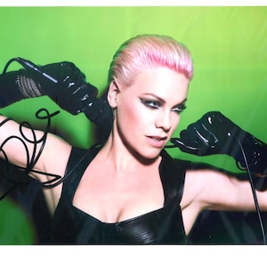 Könnte beinhalten: Ein Porträt einer Person mit rosa Haaren und dunklem Augen-Make-up, die zwei schwarze Mikrofone hält. Die Person trägt ein schwarzes Oberteil und schwarze Handschuhe vor grünem Hintergrund. Das Bild ist eine Fotografie.