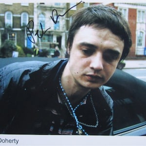 Könnte beinhalten: Ein Schwarzweißfoto eines Mannes mit dunklen Haaren und einer schwarzen Jacke. Er trägt eine blaue Perlenkette und ein schwarzes Hemd. Das Foto ist mit "Pete Doherty" signiert.