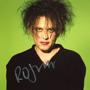 May include: A portrait of a person with messy dark hair, dark eye makeup, and dark lipstick, wearing a black V-neck sweater against a bright green background. A signature in silver ink is visible across the lower left side of the image.