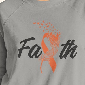 Pode incluir: Uma camisa de moletom cinza com a palavra "Faith" impressa em preto. Uma fita laranja com um design de pena está atrás da palavra "Faith". Um rastro de pequenas borboletas laranja está acima da fita.