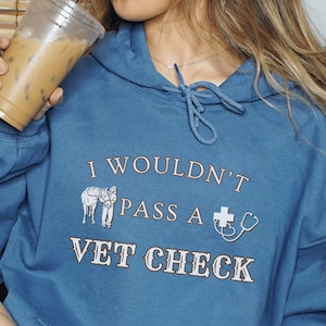 Peut inclure: Un sweat-shirt à capuche bleu avec le texte "I wouldn't pass a vet check" et un graphique de deux ânes. Le modèle porte le sweat-shirt et une casquette de baseball bleu marine.