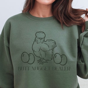 Peut inclure: Un sweat-shirt vert avec une illustration en noir et blanc d'une poule avec trois œufs et le texte "Butt Nugget Dealer".