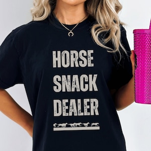 Peut inclure: T-shirt noir avec le texte "HORSE SNACK DEALER" en blanc avec une rangée de chevaux courant sous le texte.