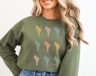 Puede incluir: Una sudadera verde con cuello redondo y mangas largas presenta un patrón de nueve siluetas de unicornios de colores. La sudadera la lleva puesta una persona con el pelo largo rubio y unos vaqueros azules.