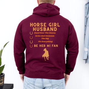Peut inclure: Un sweat à capuche bordeaux avec un graphique de fer à cheval doré et le texte "HORSE GIRL HUSBAND" avec une liste de tâches : "Hand Over The Money", "Drive And Maintain The Rig", "Fix Everything", et "BE HER #1 FAN".