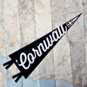 May include: Black pennant with white text that reads "Cornwall" on a white background. The pennant is folded and placed on a map.