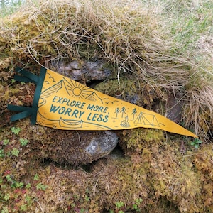 May include: A yellow pennant with green trim and the text "Explore More, Worry Less". The pennant features a campfire, trees, and a tent.