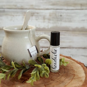 May include: A clear glass roll-on bottle with a black cap and the word "headache" in cursive script. Beside it is a beige mug with a tea bag and a spoon, and some green leaves, all on a wooden slice.