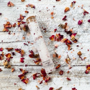 May include: A glass tube filled with white bath salts and rose petals. The tube has a cork top and a label that reads "Bath Salts Rose". The tube is surrounded by rose petals and pink Himalayan salt crystals on a white wooden surface.