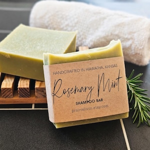 May include: Two green Rosemary Mint shampoo bars, one on a wooden soap dish, the other wrapped in a brown paper label. The label reads "Handcrafted in Hiawatha, Kansas" and "prairielivin.etsy.com". A white towel and rosemary sprig are in the background.