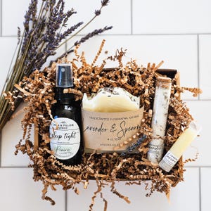 May include: A gift box filled with lavender-themed bath and body products. The box contains a lavender and spearmint soap, a linen and pillow spray labeled "sleep tight", a tube of lavender lip balm, and a small glass jar of lavender bath salts.