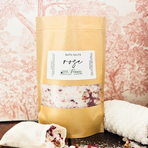 May include: A brown paper bag filled with white bath salts and dried rose petals. The bag has a label that reads "BATH SALTS rose Prairie LIVIN".
