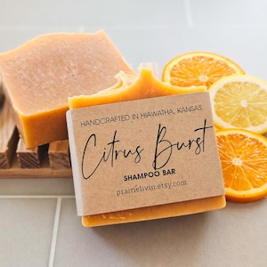 May include: A close-up of a citrus-scented shampoo bar with a kraft paper label that reads "Citrus Burst" and "Shampoo Bar". The bar is orange and is next to orange and lemon slices. The text "Handcrafted in Hiawatha, Kansas" is also on the label.
