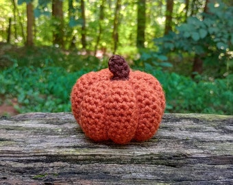 May include: A crocheted orange pumpkin with a brown stem, sitting on a weathered wooden log.