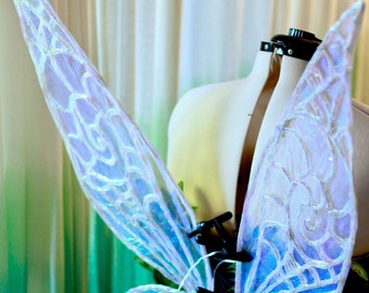 Puede incluir: Un par de alas de hada azul iridiscente con un patrón blanco. Las alas están unidas a un marco de alambre negro y se muestran en un maniquí blanco.