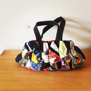 Puede incluir: Un bolso tote de patchwork colorido con correas negras. El bolso está hecho de diferentes telas en varios colores y patrones, incluyendo rojo, azul, amarillo y verde.