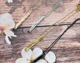 May include: Four minimalist bar necklaces in gold, silver, and black. Each necklace features a slender rectangular pendant suspended from a delicate chain. The necklaces are displayed on a wooden surface with white flower accents.
