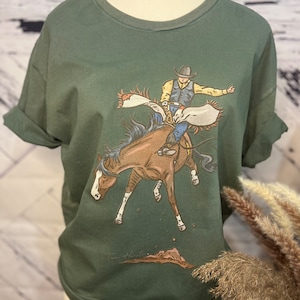 Puede incluir: Una camiseta verde con un gráfico de un vaquero montando un caballo que se resiste. El gráfico está en tonos de marrón, amarillo y azul. El texto "Tim Hunt" está impreso en la parte inferior de la camiseta.