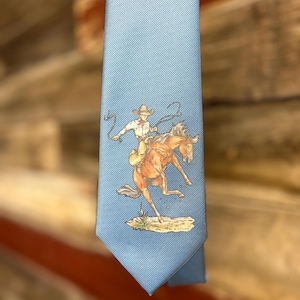 May include: A light blue necktie featuring a detailed illustration of a cowboy riding a bucking brown horse while swinging a lasso. The rider wears a hat and light-colored clothing, and the scene is set on a small patch of ground with grass.
