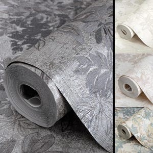 May include: A roll of wallpaper featuring a metallic silver floral pattern on a textured dark gray background, alongside three additional rolls in lighter color variations including cream, beige, and a soft blue and tan floral design.