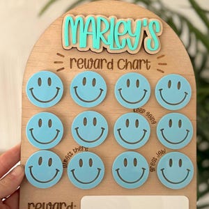 May include: Wooden reward chart with the name "Marley's" in turquoise letters. The chart features twelve light blue smiley face tokens and the words "reward chart" and "reward:" in a dark brown font.