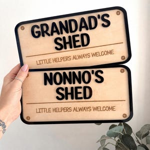May include: Two rectangular signs with light wood backgrounds and black borders. The top sign reads GRANDAD'S SHED and the bottom sign reads NONNO'S SHED. Both signs feature the text LITTLE HELPERS ALWAYS WELCOME and four decorative screw heads.