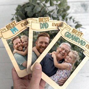 May include: Wooden photo frames with the phrases "Best Pa Ever", "Best Dad Ever", and "Best Grandad Ever" in a banner design. Each frame holds a photo of a person with a child. The frames are held in a hand.