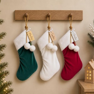 May include: Three velvet Christmas stockings in dark green, white, and red, each featuring a white faux fur cuff, two white pom-poms, and a wooden name tag labelled Marley, Sophie, or Chester, hanging from a wooden wall-mounted rack.