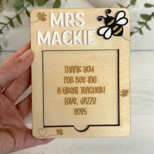 May include: A wooden plaque with the name "MRS MACKIE" in white letters and a cartoon bee. The plaque includes the message "THANK YOU for Bee-ing A GREAT TEACHER! LOVE, JAZZY 2025" in a wooden frame.