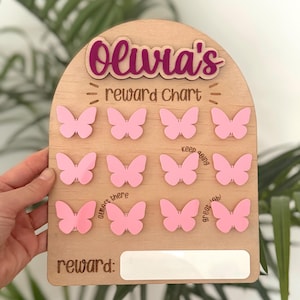 May include: Wooden reward chart featuring pink butterfly cutouts and the name "Olivia's". The chart includes the words "reward chart" and "reward:" with space for a reward to be written.