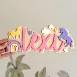 May include: A wooden wall sign featuring the name lexie in pink script lettering. The sign is decorated with a yellow unicorn, a purple unicorn, a rainbow with pink, purple, and yellow arches, white clouds, and small yellow and purple stars.
