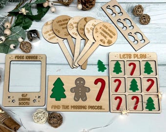 May include: A selection of wooden Christmas decorations and games. Features a photo frame with "Free Kisses" text, game pieces, and signs with phrases like "Let's Watch a Christmas Movie." The items are arranged on a light wood surface.