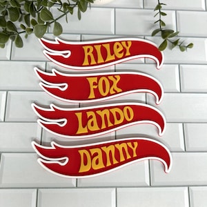 May include: Four red and white flame-shaped custom name signs featuring yellow lettering. The names displayed are Riley, Fox, Lando, and Danny. Each sign has a distinct stylised font and a white border, arranged in a vertical stack.