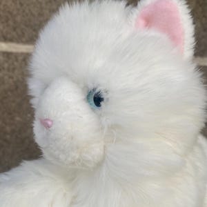 May include: White plush cat toy with blue eyes and pink ears. The cat is sitting with its head tilted to the side.