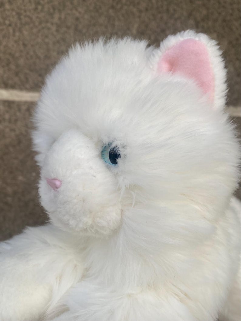 May include: White plush cat toy with blue eyes and pink ears. The cat is sitting with its head tilted to the side.